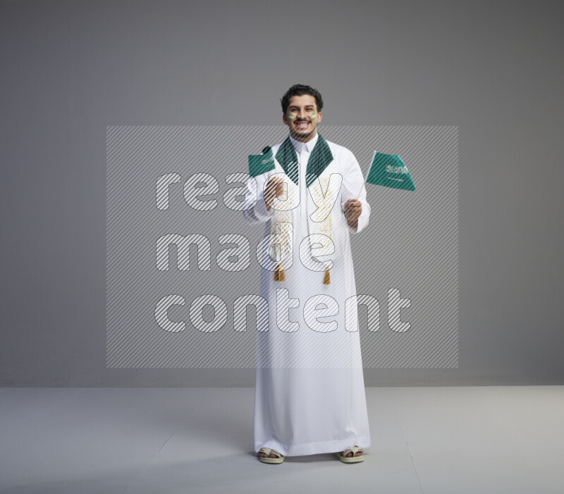 A Saudi man standing wearing thob and saudi flag scarf with face painting holding small Saudi flag on gray background