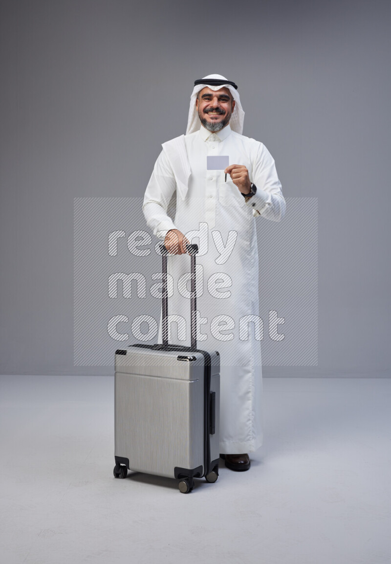 Saudi man wearing Thob and white Shomag standing holding Travel bag and ATM card on Gray background