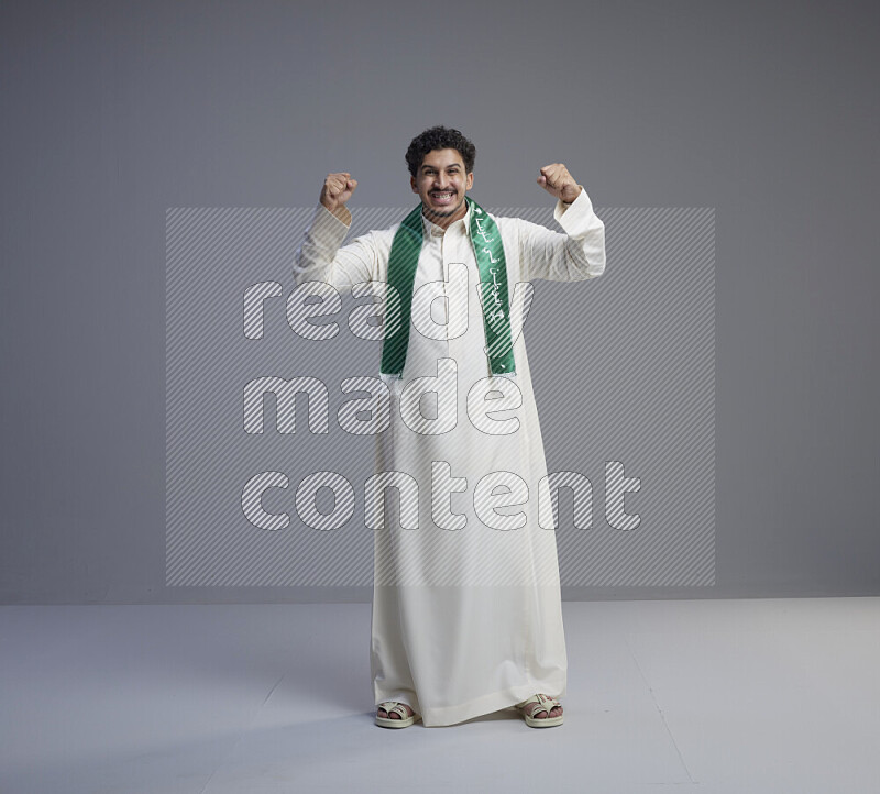 A Saudi man standing wearing thob and Saudi flag scarf on gray background