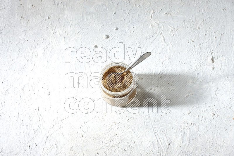 A glass jar and a metal spoon full of cumin powder on textured white flooring