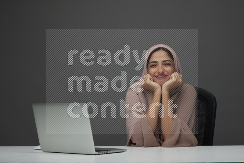 A Saudi woman Setting on her desk on a Gray Background wearing Brown Abaya with Hijab
