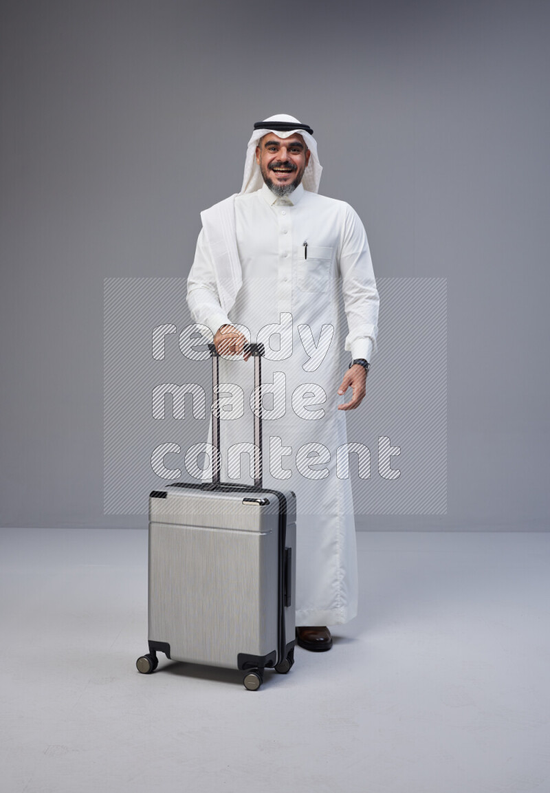 Saudi man wearing Thob and white Shomag standing holding Travel bag on Gray background