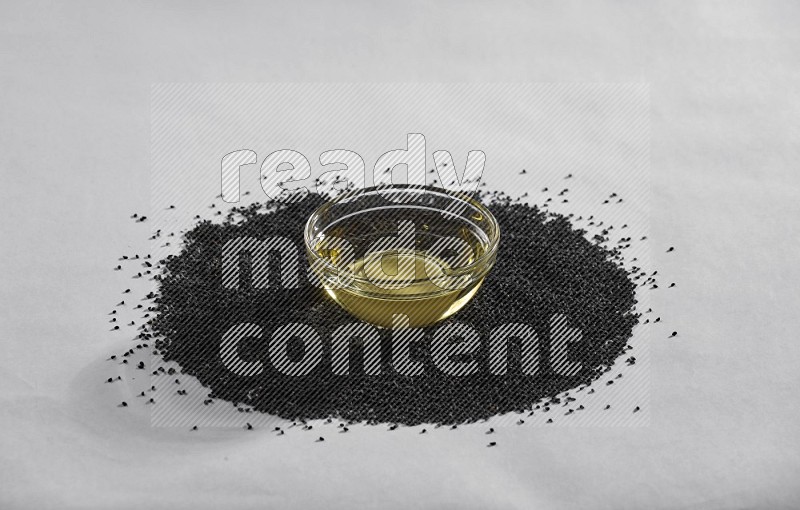 A glass bowl full of black seeds oil surrounded by the seeds on a white flooring