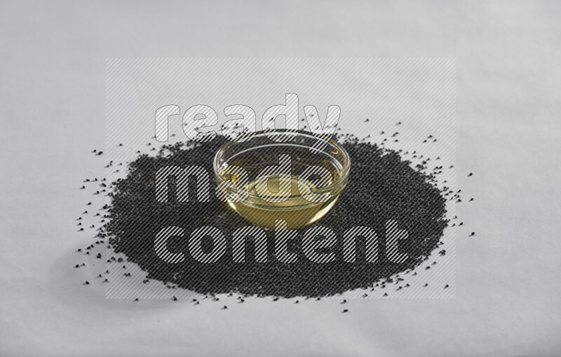 A glass bowl full of black seeds oil surrounded by the seeds on a white flooring