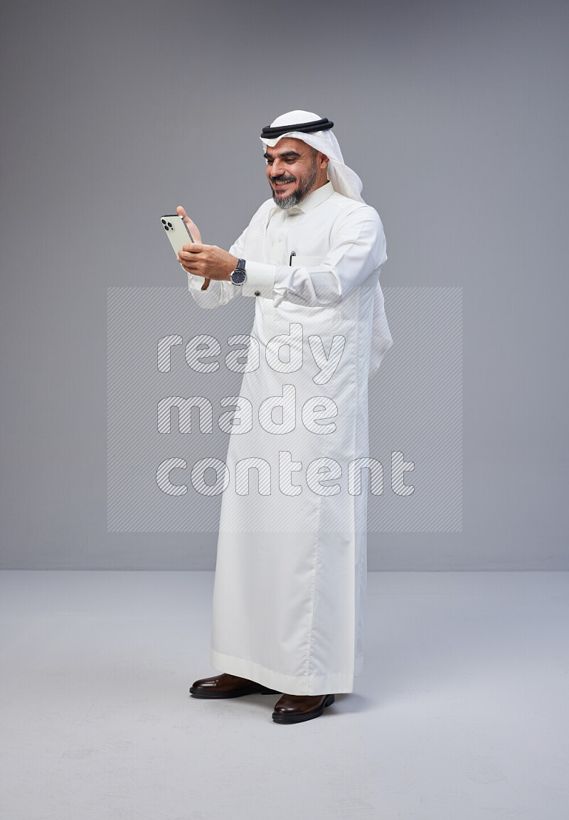 Saudi man Wearing Thob and white Shomag standing texting on phone on Gray background