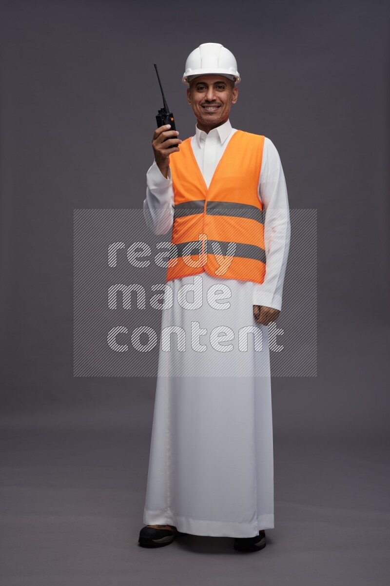 Saudi man wearing thob with engineer vest standing holding walkie-talkie on gray background