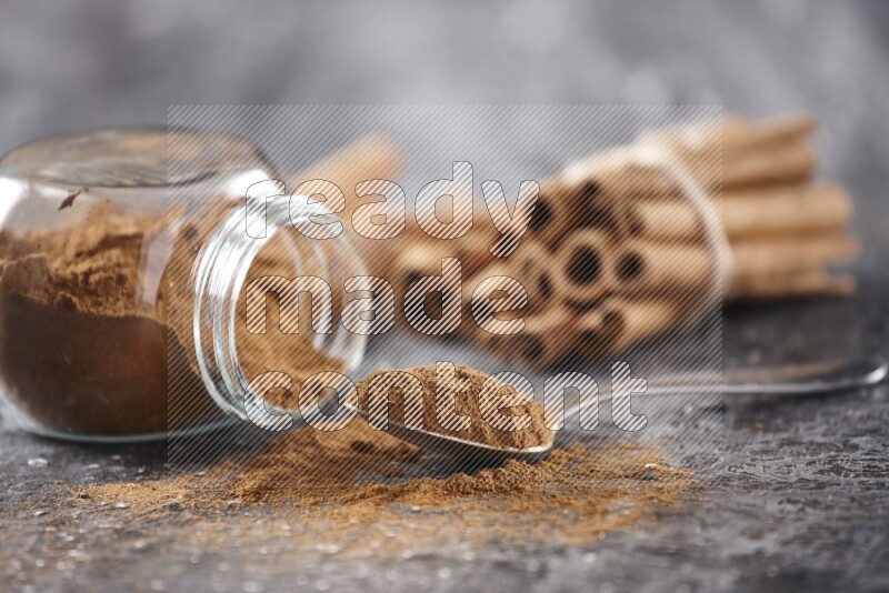 Herbal glass jar full cinnamon powder flipped and a metal spoon full of powder, cinnamon sticks stacked and bounded in the back on textured black background in different angles