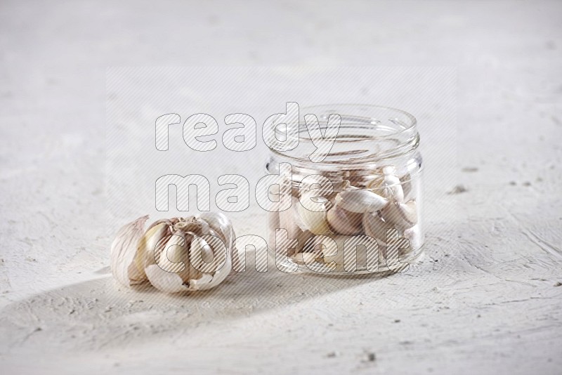 A glass jar full of garlic cloves and beside it a garlic bulb on a textured white flooring in different angles