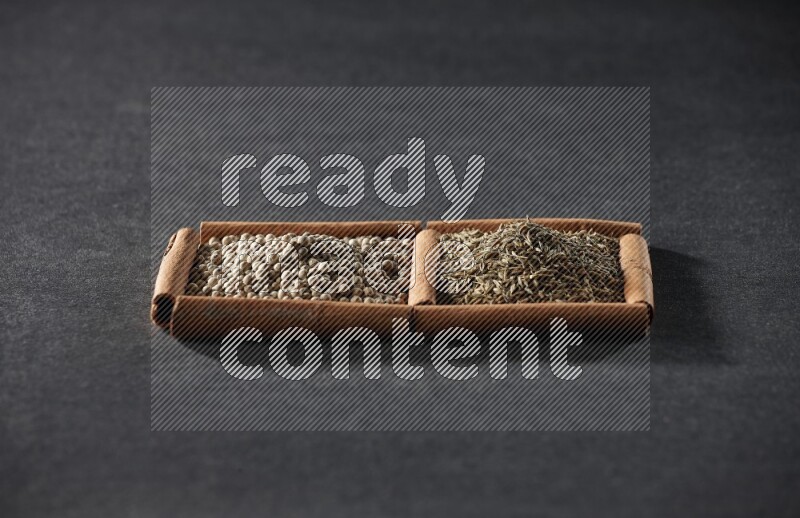 مربعان من أعواد القرفة ممتلئان بحبوب الفلفل الأبيض وحبوب الكمون علي خلفية سوداء