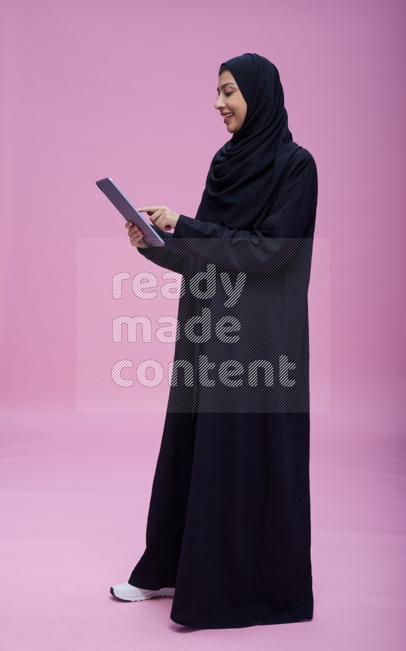 Saudi woman wearing Abaya standing working on tablet on pink background