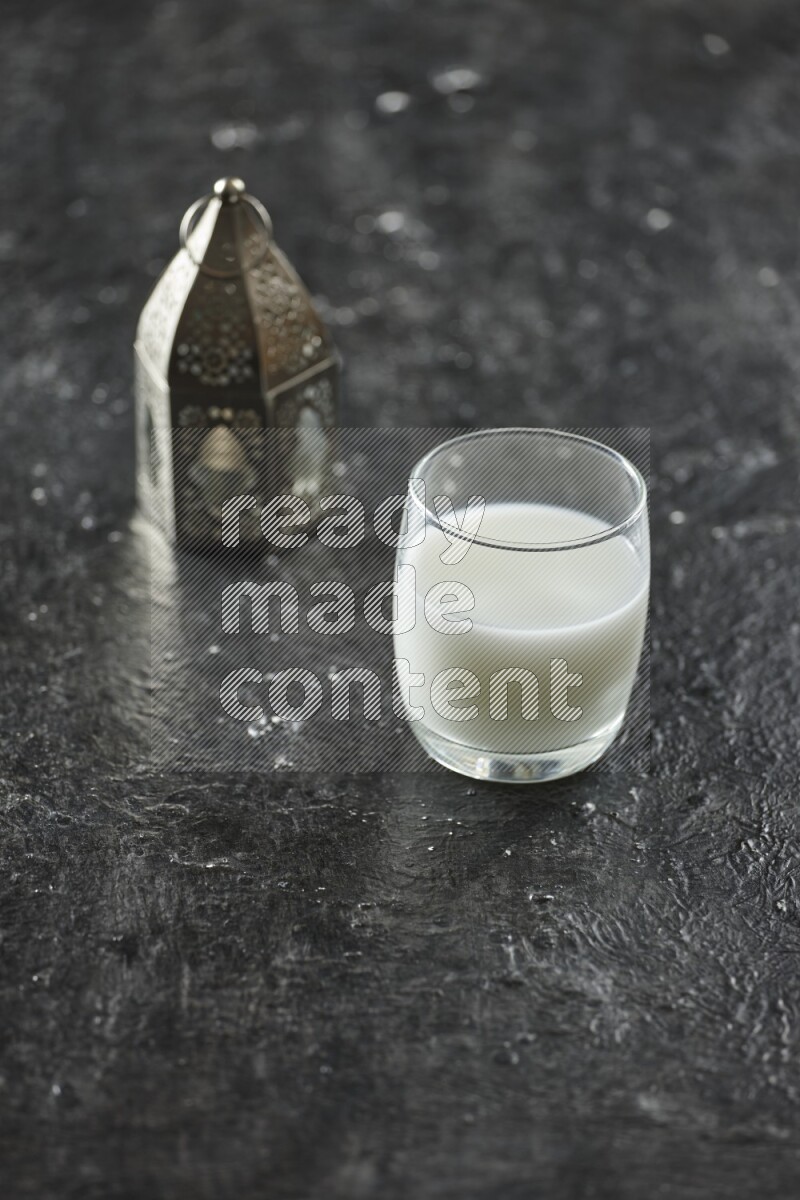 A silver lantern with different drinks, dates, nuts, prayer beads and quran on textured black background