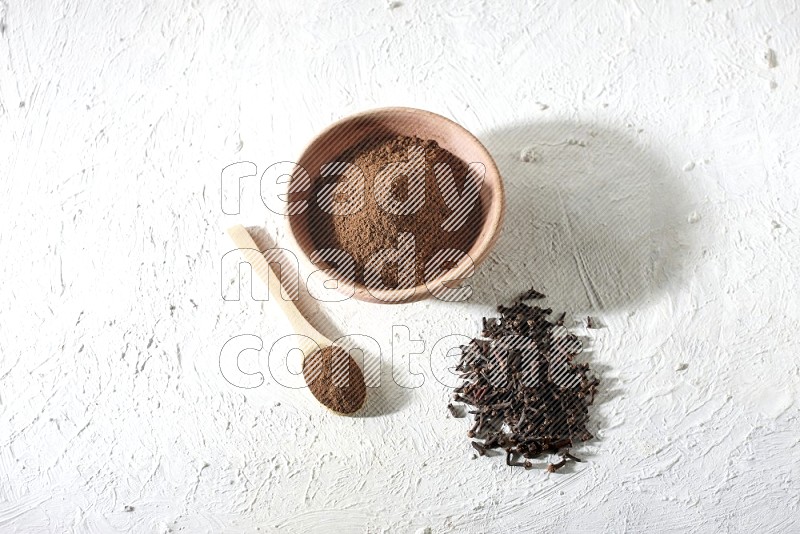 A wooden bowl and wooden spoon full of cloves powder with cloves spread on textured white flooring