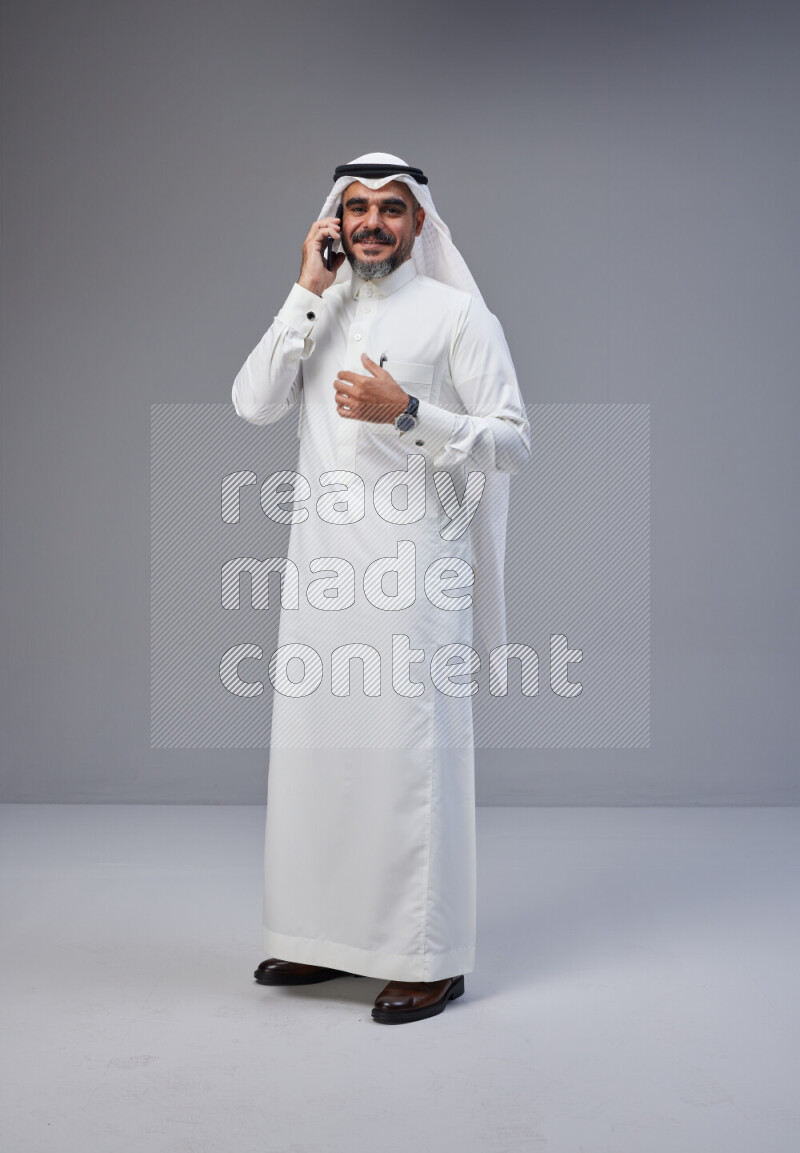 Saudi man Wearing Thob and white Shomag standing talking on phone on Gray background