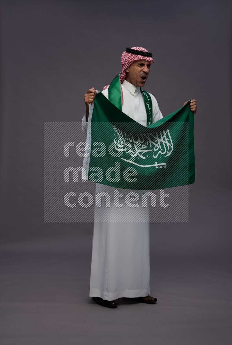 Saudi man wearing thob and shomag standing holding Saudi flag on gray background