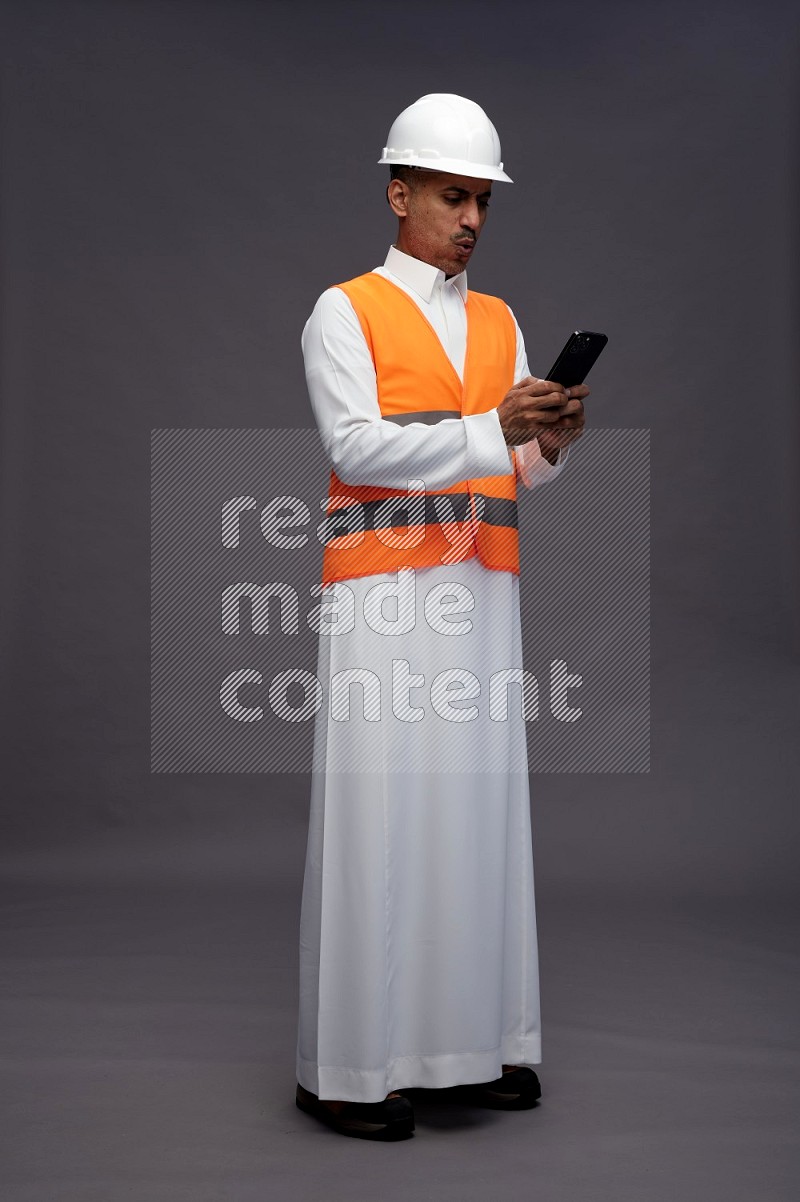 Saudi man wearing thob with engineer vest standing texting on phone on gray background