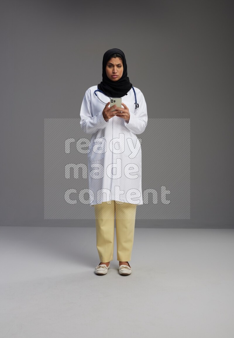 Saudi woman wearing lab coat with stethoscope standing texting on phone on Gray background