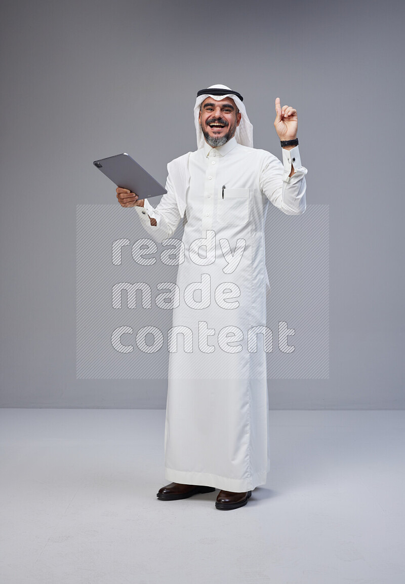 Saudi man Wearing Thob and white Shomag standing working on tablet on Gray background