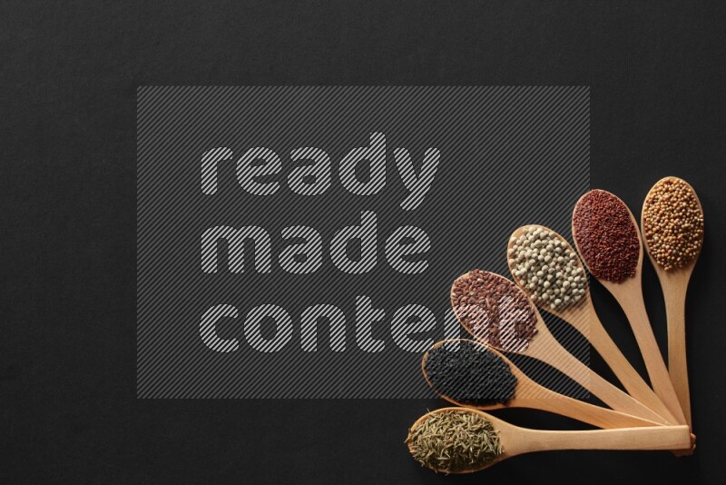 wooden spoons filled with white peppers, mustard seeds, black seeds, garden cress, cumin and flax on black flooring