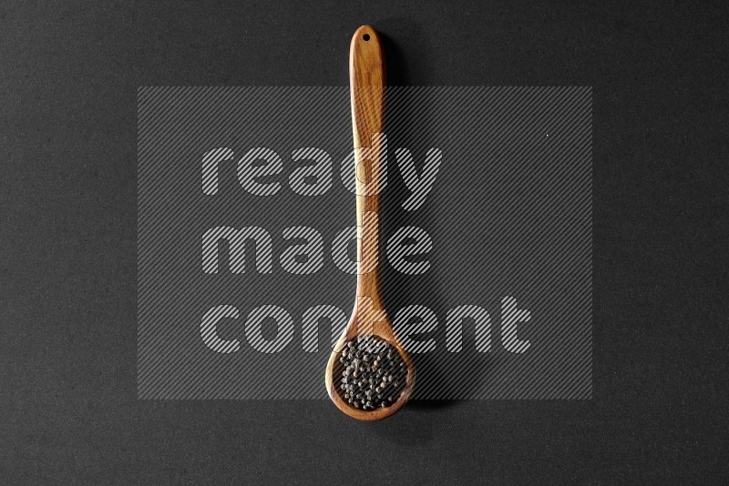 A wooden ladle full of black pepper beads on a black flooring