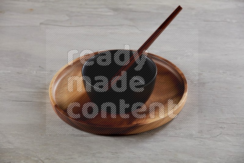 black ceramic round bowl on top of brown wood round plate and wood chopsticks, on grey textured countertop