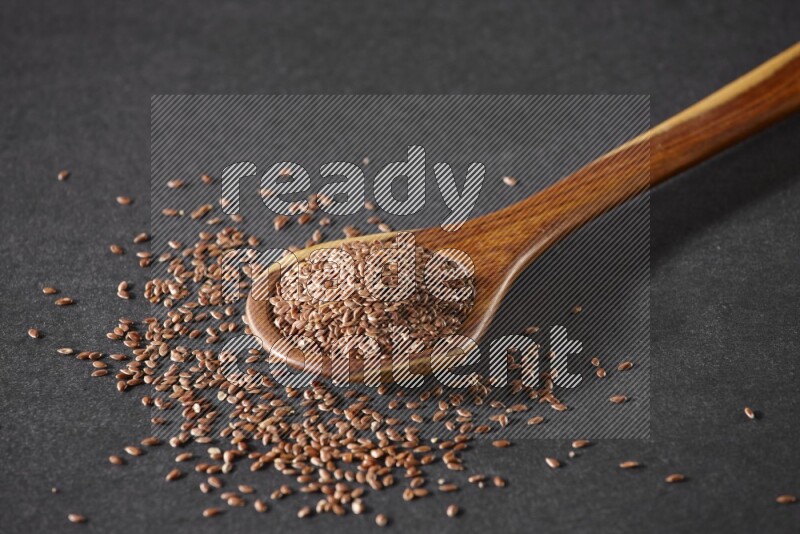 A wooden ladle full of flaxseeds and seeds spread beside it on a black flooring