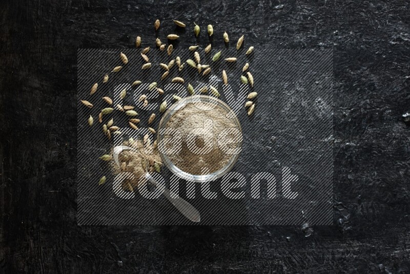 وعاء زجاجي وملعقة معدنية ممتلئان ببودرة الهيل مع حبوب الهيل بجانبهما على خلفية سوداء