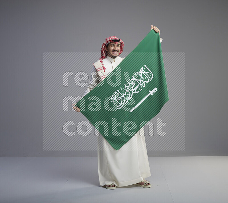 A saudi man standing wearing thob and red shomag holding big saudi flag on gray background