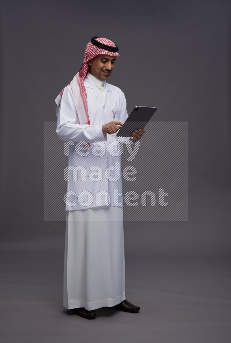 Saudi man wearing thob with lab coat and shomag with pocket employee badge standing working on tablet on gray background