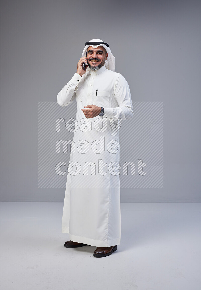 Saudi man Wearing Thob and white Shomag standing talking on phone on Gray background