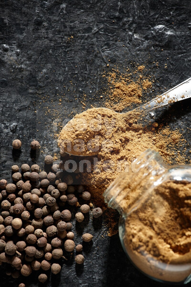 A flipped glass spice jar and metal spoon full of allspice powder and powder spilled out of it with whole balls on a textured black flooring