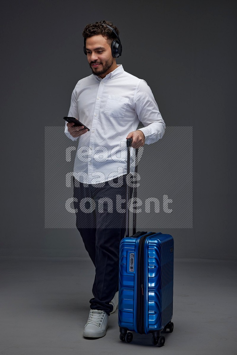 A man wearing smart casual using his phone while wearing headphone and holding luggage eye level on a gray background