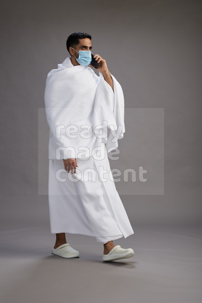 A man wearing Ehram with face mask Standing talking on phone on gray background