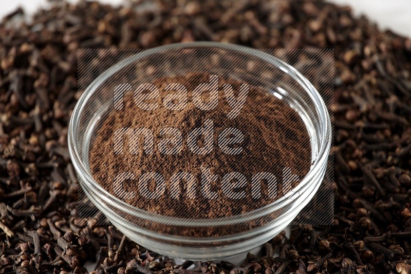 A glass bowl full of cloves powder and cloves grains spread on white flooring