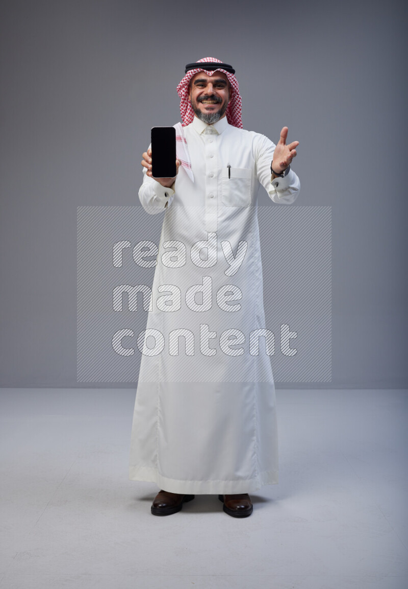Saudi man Wearing Thob and red Shomag standing showing phone to camera on Gray background