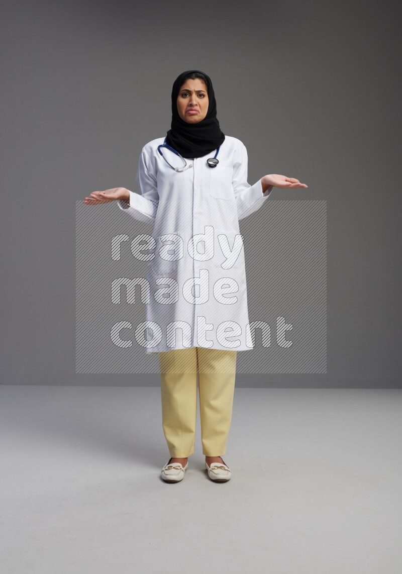 Saudi woman wearing lab coat with stethoscope standing interacting with the camera on Gray background