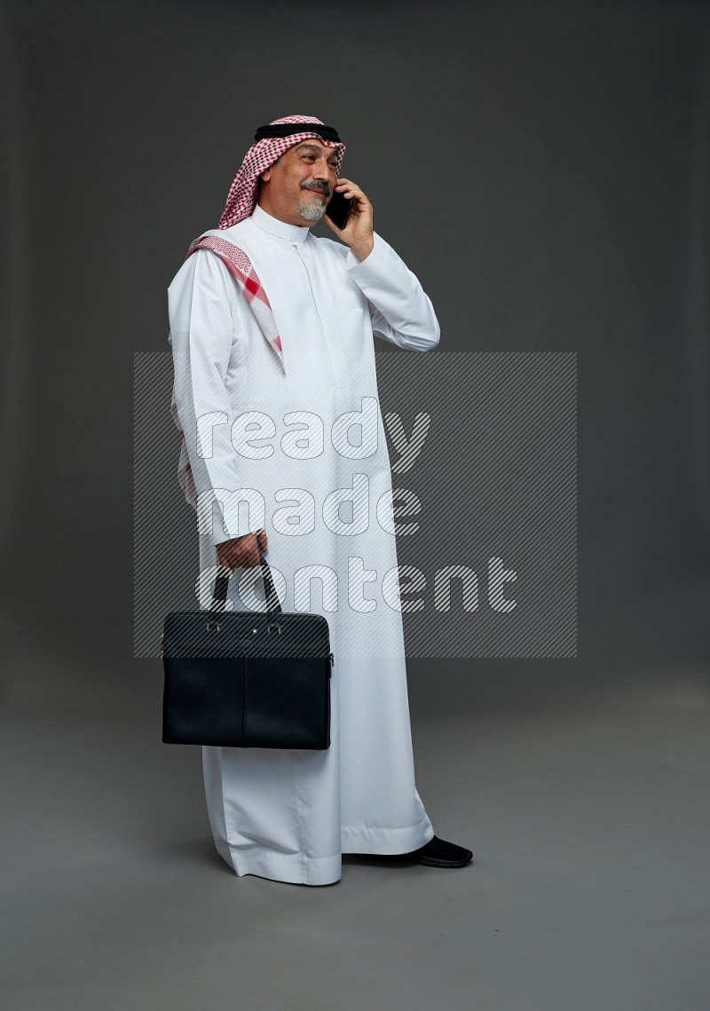 Saudi man with shomag Standing holding bag talking on phone on gray background