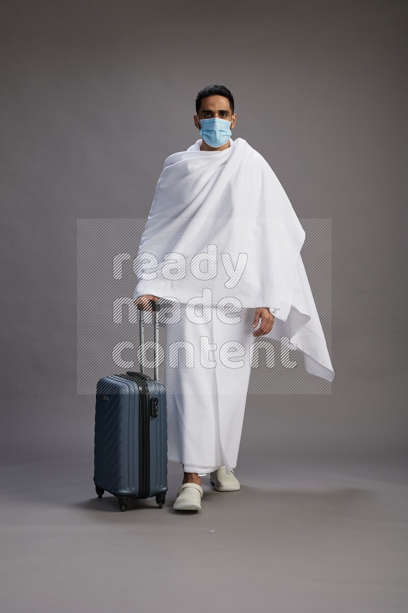 A man wearing Ehram with face mask Standing holding traveling bag on gray background