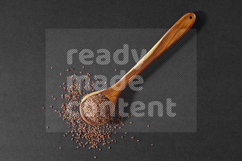 A wooden ladle full of flaxseeds and seeds spread beside it on a black flooring