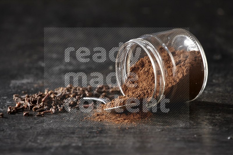 A flipped glass jar and metal spoon full of cloves powder with cloves spread on a textured black flooring