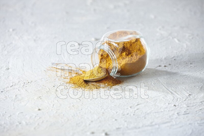 A flipped glass spice jar and a metal spoon full of turmeric powder and powder spilled out of it on textured white flooring
