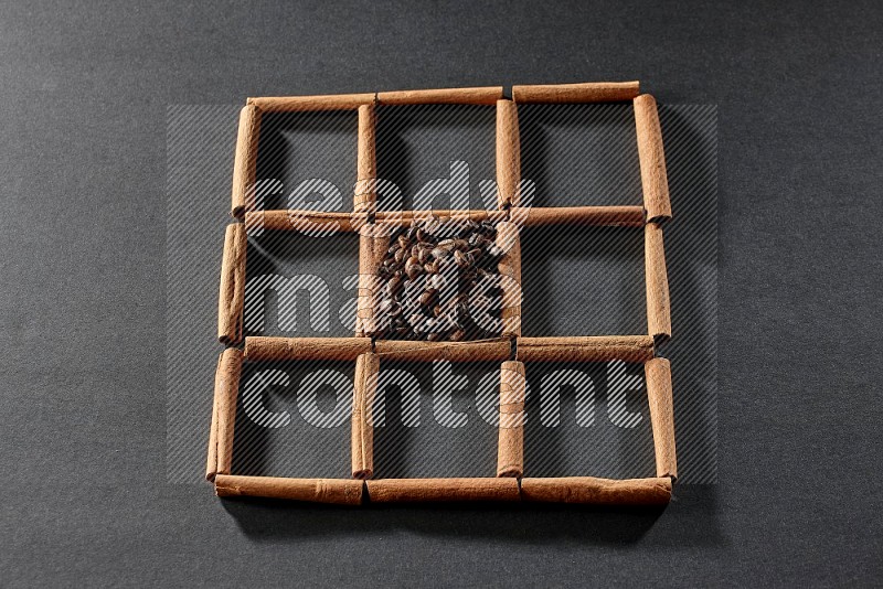9 squares of cinnamon sticks full of coffee beans in the middle surrounded by nutmeg, dried mint, cloves, dried basil, dried ginger, cinnamon, star anise and cardamom on black flooring