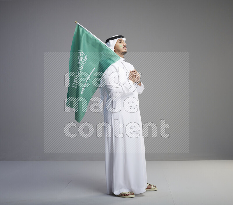 A Saudi man standing wearing thob and white shomag with face painting raising big Saudi flag on gray background