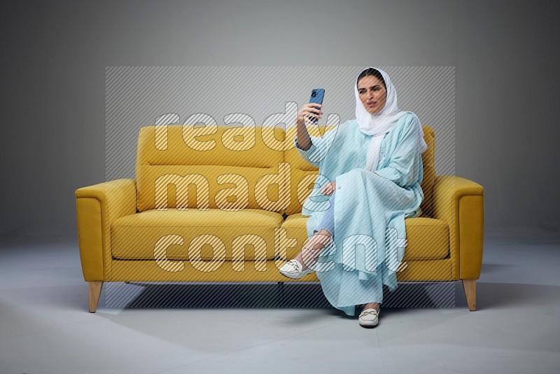 A Saudi woman wearing a light blue Abaya and white head scarf sitting on a yellow sofa and taking selfies with her phone eye level on a grey background