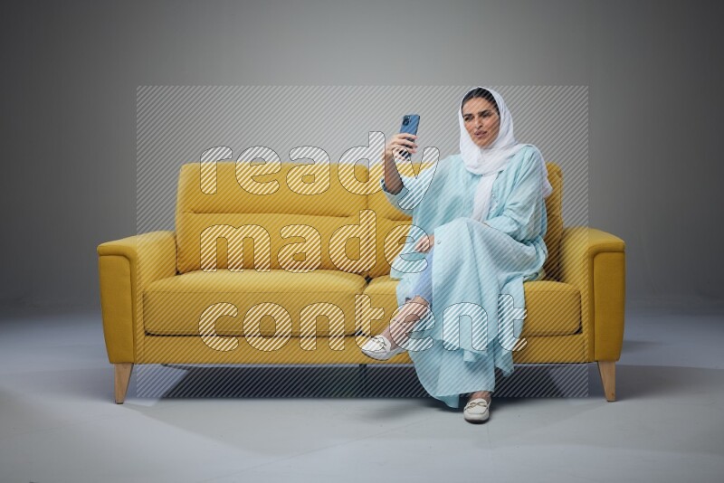 A Saudi woman wearing a light blue Abaya and white head scarf sitting on a yellow sofa and taking selfies with her phone eye level on a grey background