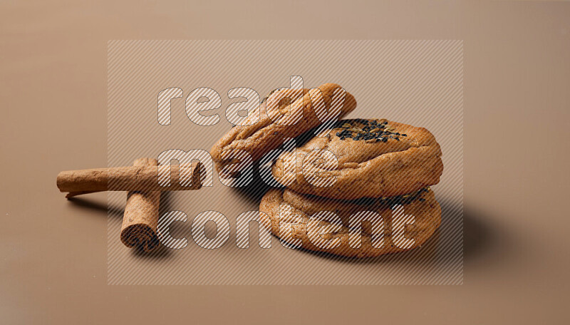 Three chocolate chip cookies beside cinimon sticks on a brown background