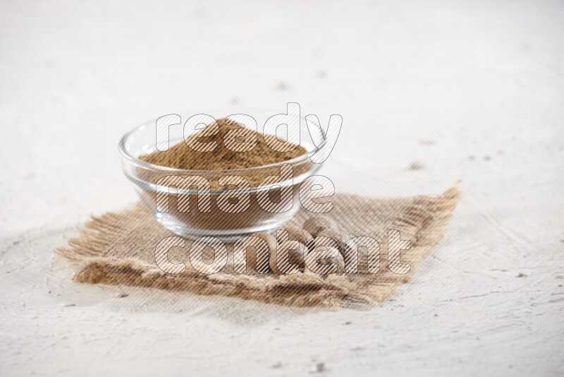 A glass bowl full of nutmeg powder with whole seeds beside it on burlap fabric on a textured white flooring