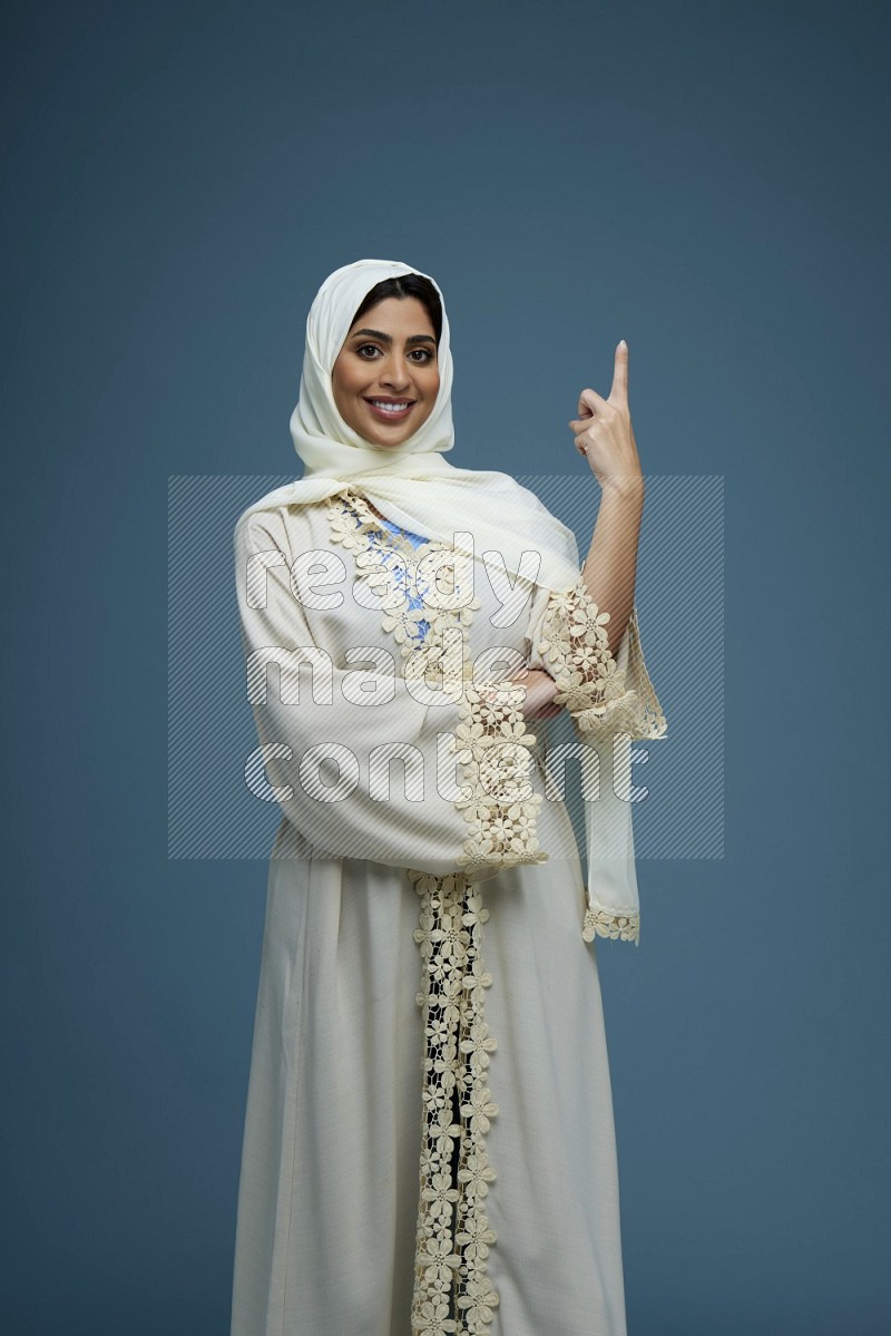 A Saudi woman pointing in a blue background wearing an off-white Abaya Hijab