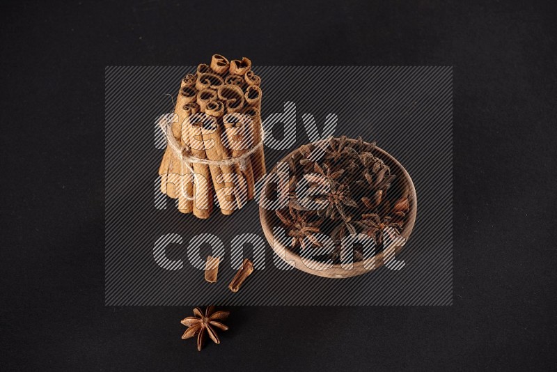 A stacked and bounded cinnamon sticks and a wooden bowl full of star anise on a black background