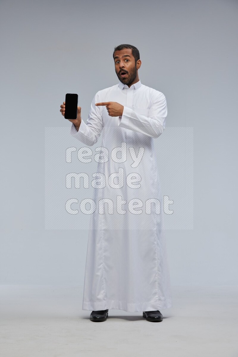 Saudi man wearing Thob standing showing phone to camera on Gray background