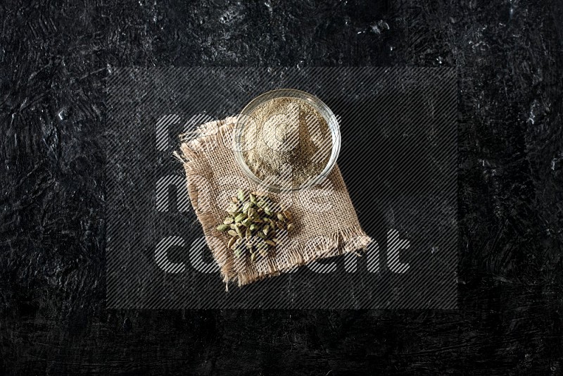 A glass bowl full of cardamom powder with cardamom seeds on a burlap piece on textured black flooring